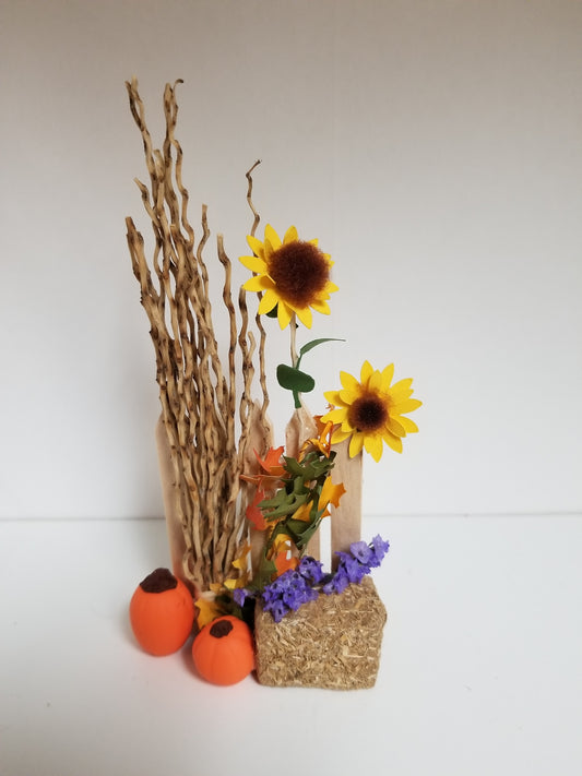 Miniature wooden fence with bale of hay, corn stalks, pumpkins, sunflowers, and colored leaves.